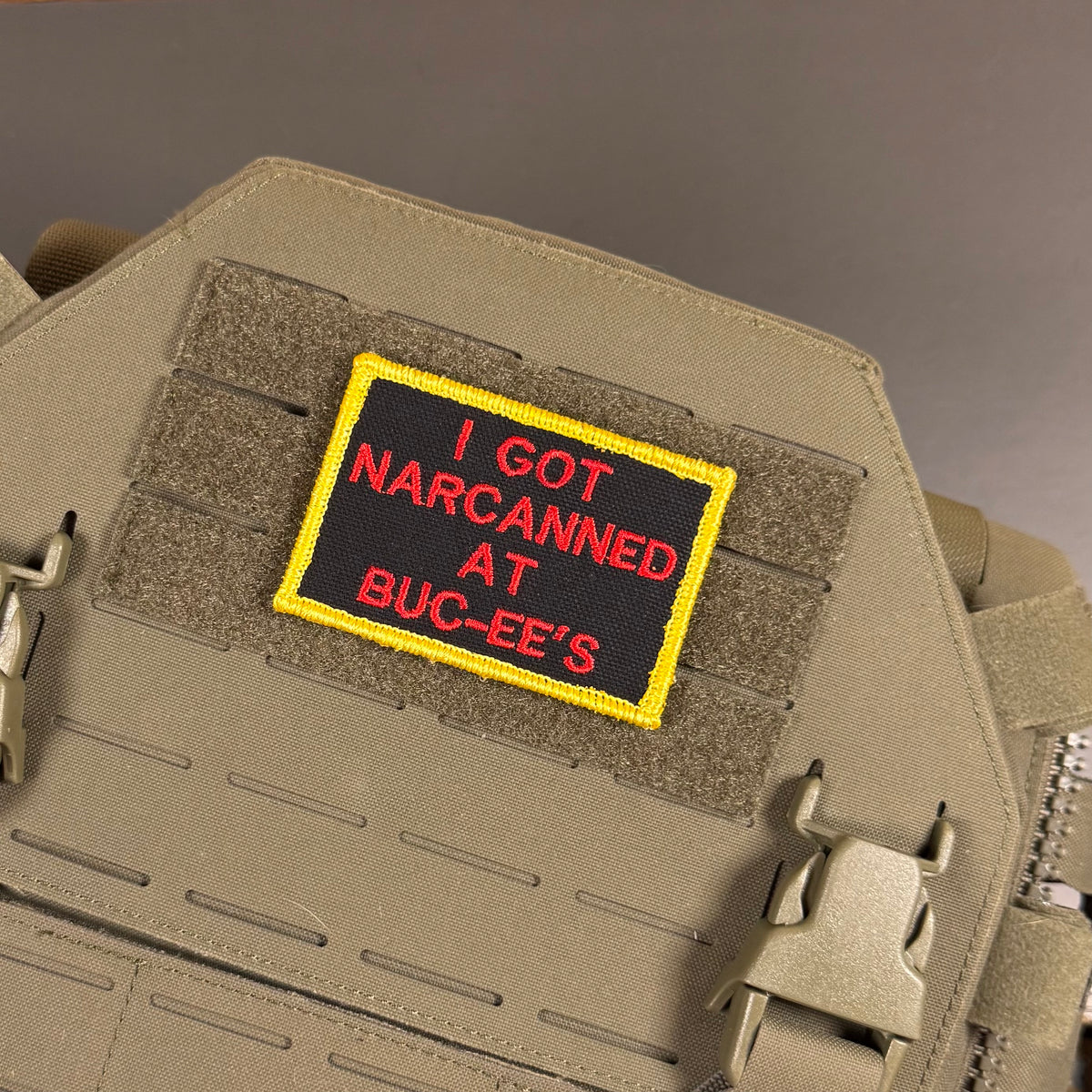 A rectangular embroidered patch with a black background, yellow border, and red text reading 'I GOT NARCANED AT BUC-EE'S', attached to tan tactical velcro panels on a tan vest or plate carrier.