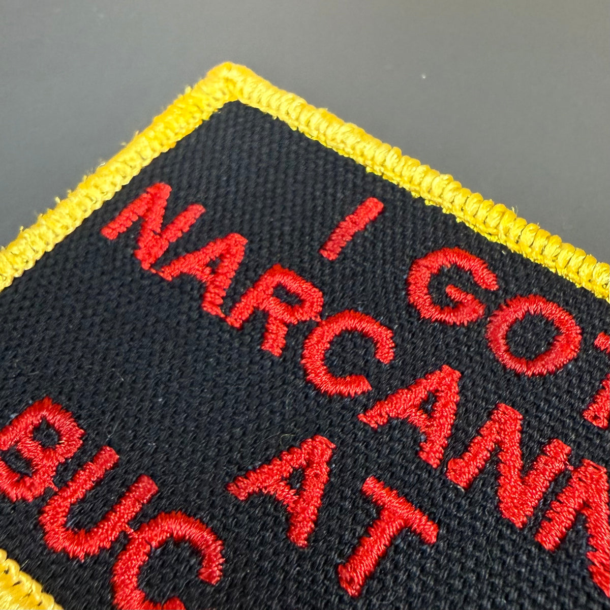 Close-up of a rectangular embroidered patch with a black field, bright yellow border, and red text reading 'I GOT NARCAN' and 'BUT AT'.