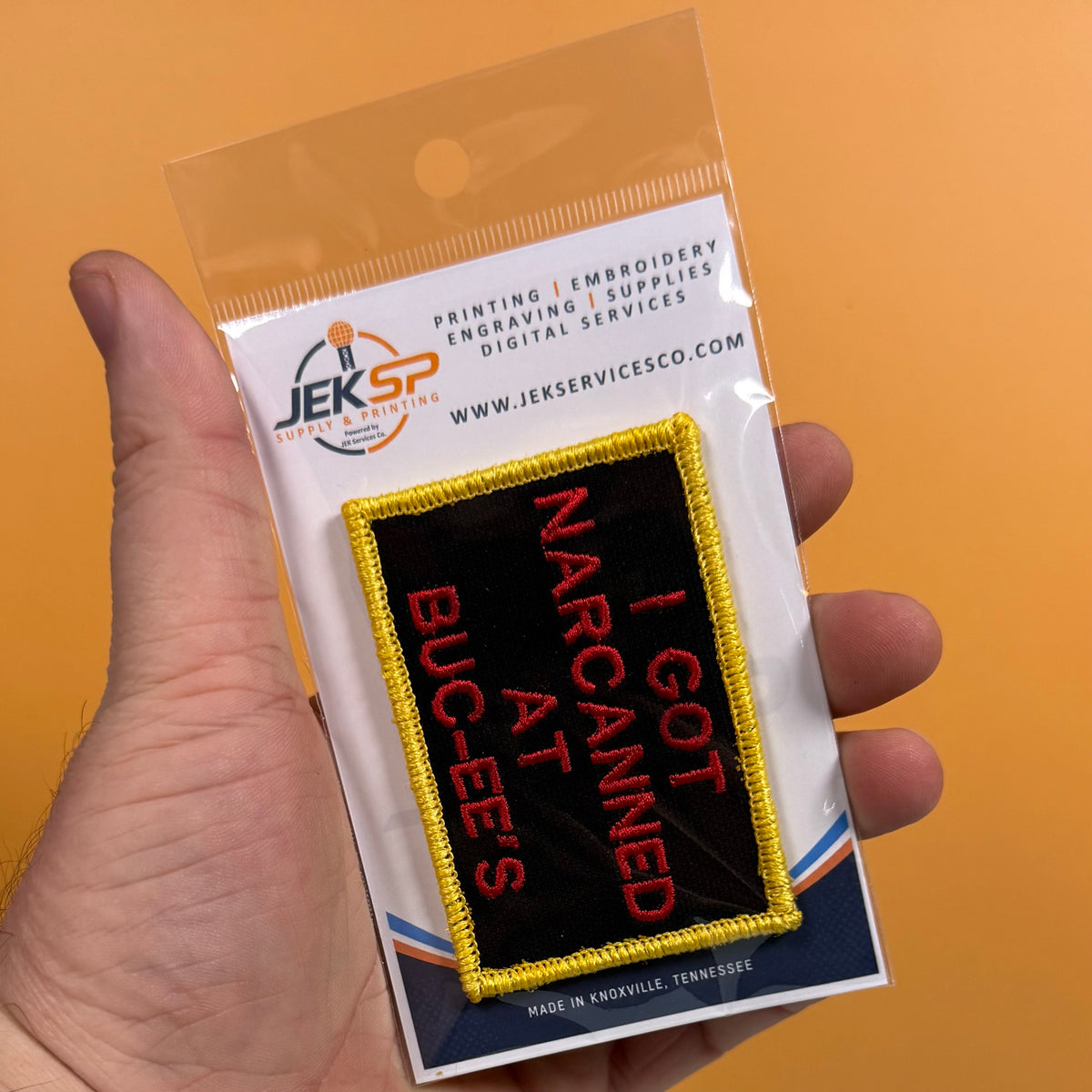 A hand holds a clear plastic blister pack containing an embroidered patch. The patch is black with a yellow border and red text reading 'I GOT NARCIAMED AT BUC-EE'S'.