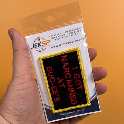 A hand holds a clear plastic blister pack containing an embroidered patch. The patch is black with a yellow border and red text reading 'I GOT NARCIAMED AT BUC-EE'S'.