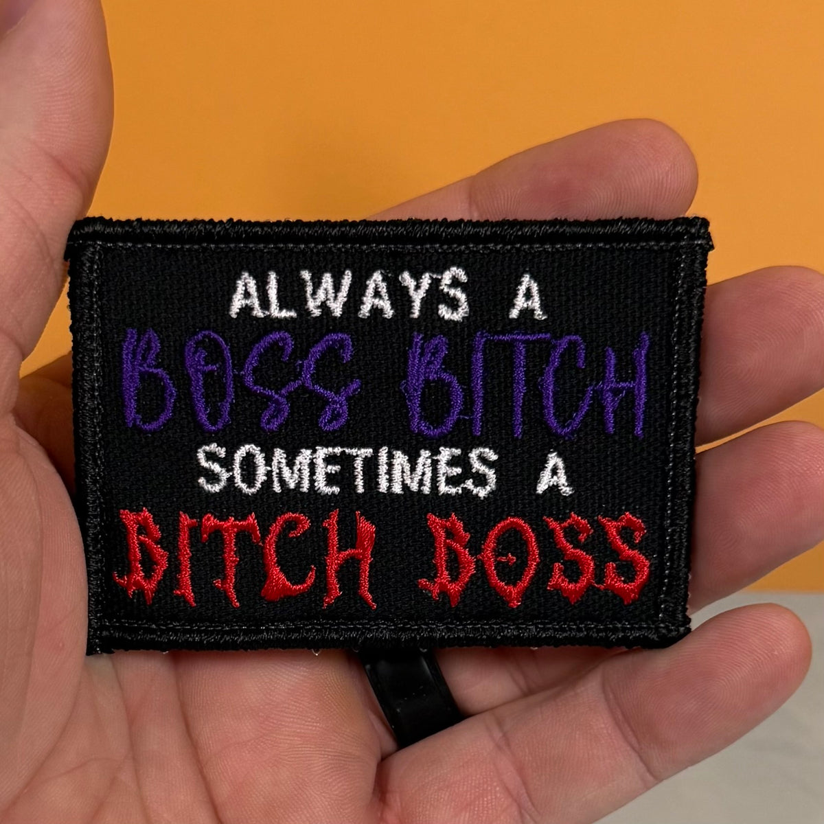 A close-up of a hand holding a rectangular embroidered patch with the text 'ALWAYS A BOSS BITCH SOMETIMES A BITCH BOSS' in white, purple, and red embroidery on a black background, against a solid orange backdrop.