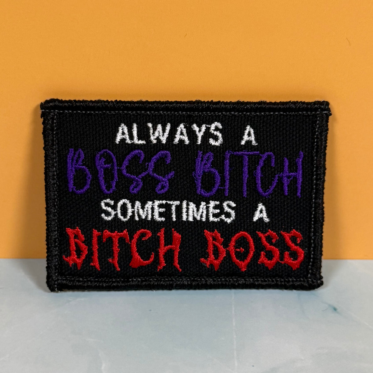 A rectangular embroidered patch with a black background and bold text reading 'ALWAYS A BOSS BITCH SOMETIMES A BITCH BOSS' in white, purple, and red lettering, displayed against a solid orange background.