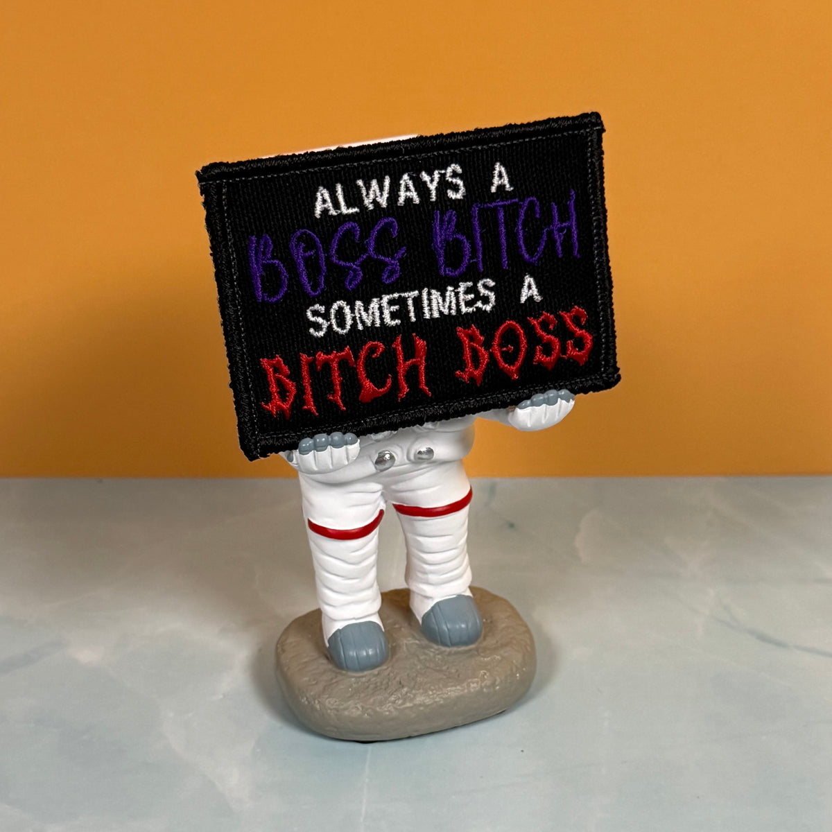 A novelty astronaut figurine holding a black embroidered sign with the text 'ALWAYS A BOSS BITCH SOMETIMES A BITCH BOSS' in white, purple, and red lettering, set against an orange background.