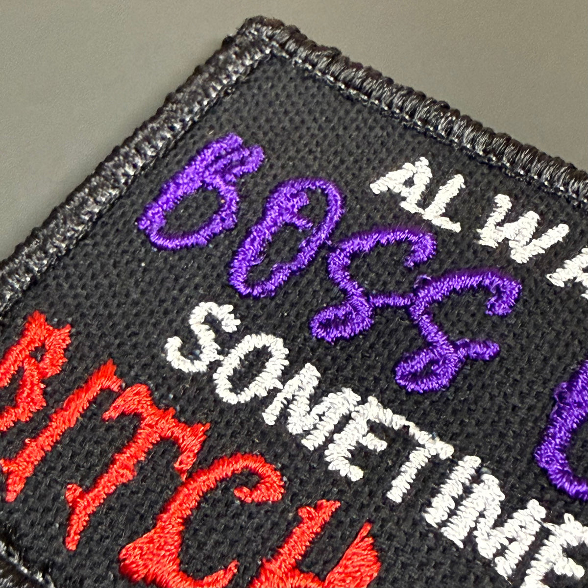 Close-up of an embroidered patch with black mesh background and text reading 'ALWAYS BOSS SOMETIMES BITCH' in purple, white, and red embroidery.