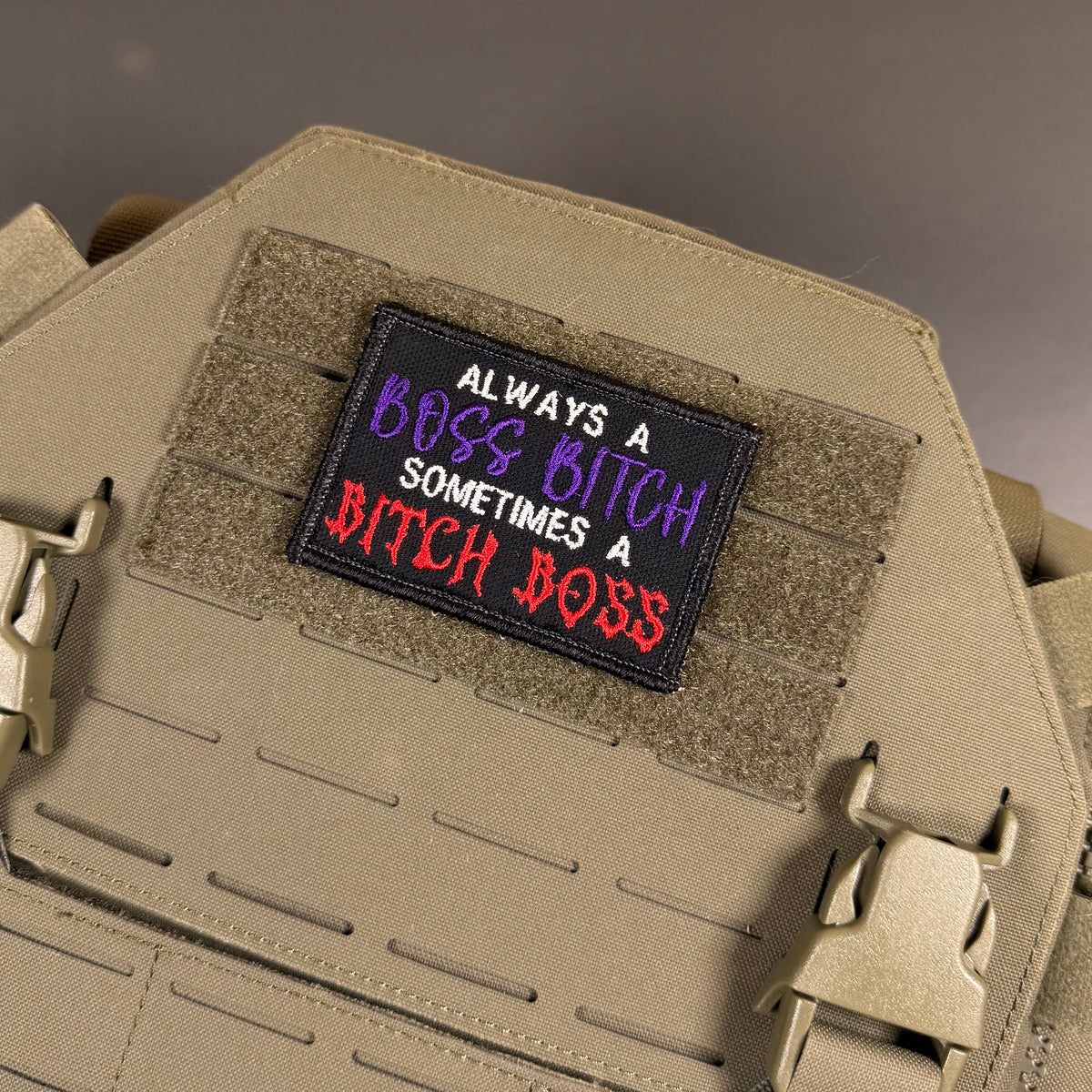 A rectangular embroidered patch with the text 'ALWAYS A BOSS BITCH SOMETIMES A BITCH BOSS' in white, purple, and red lettering on a black background, attached to a tan tactical vest with hook-and-loop panels and plastic quick-release buckles.