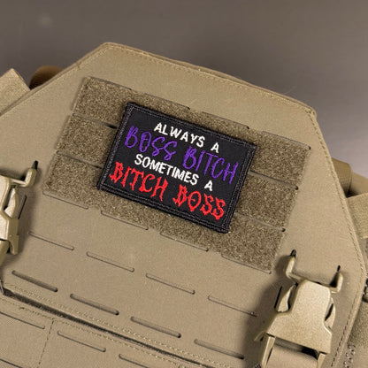 A rectangular embroidered patch with the text 'ALWAYS A BOSS BITCH SOMETIMES A BITCH BOSS' in white, purple, and red lettering on a black background, attached to a tan tactical vest with hook-and-loop panels and plastic quick-release buckles.