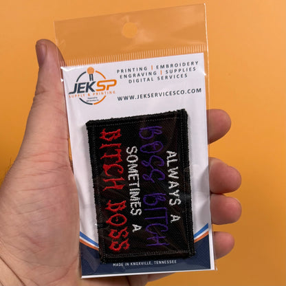 A hand holds a clear plastic bag containing an embroidered patch. The patch is black with red, purple, and white text reading 'ALWAYS BITCH SOMETIMES A BOSS'.