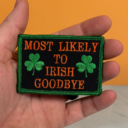 A hand holding a rectangular embroidered patch with the text 'MOST LIKELY TO IRISH GOODBYE' in orange on a black background, with green shamrocks and a green border, against an orange background.