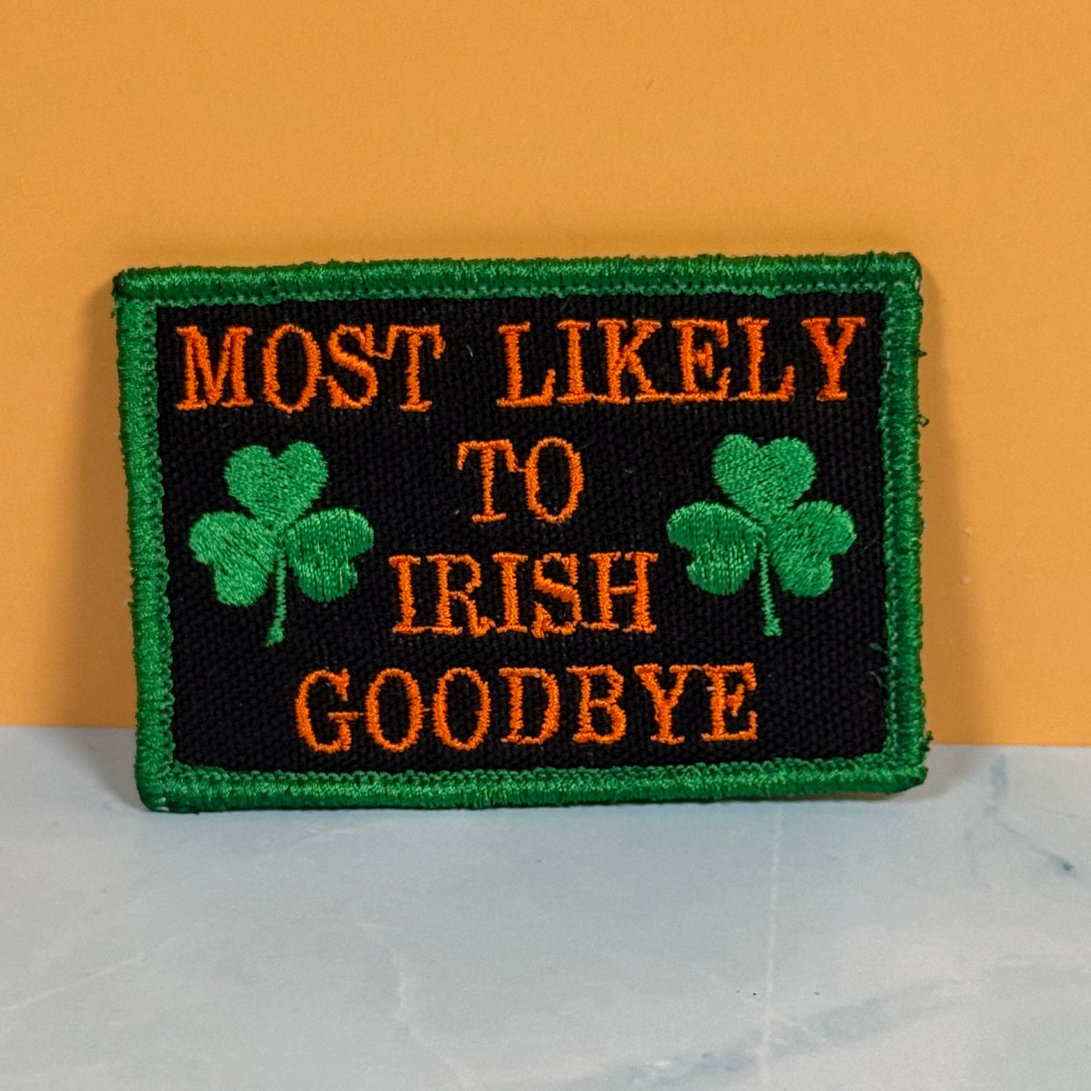 An embroidered rectangular patch with a green border, black background, orange text reading 'MOST LIKELY TO IRISH GOODBYE,' and two green shamrocks, shown against an orange wall and light marble surface.