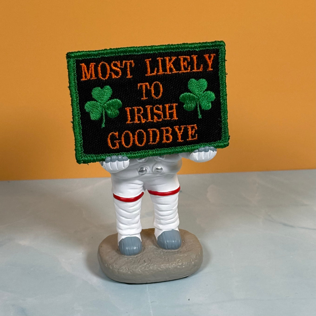 A novelty figurine of an astronaut holding a black embroidered sign that reads 'MOST LIKELY TO IRISH GOODBYE' in orange letters with green shamrocks, set against an orange background.