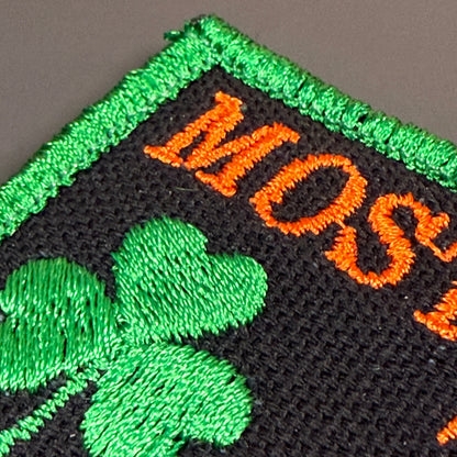 Close-up of an embroidered patch with a black mesh background, bright green border and shamrock, and orange lettering partially reading 'MOS'.