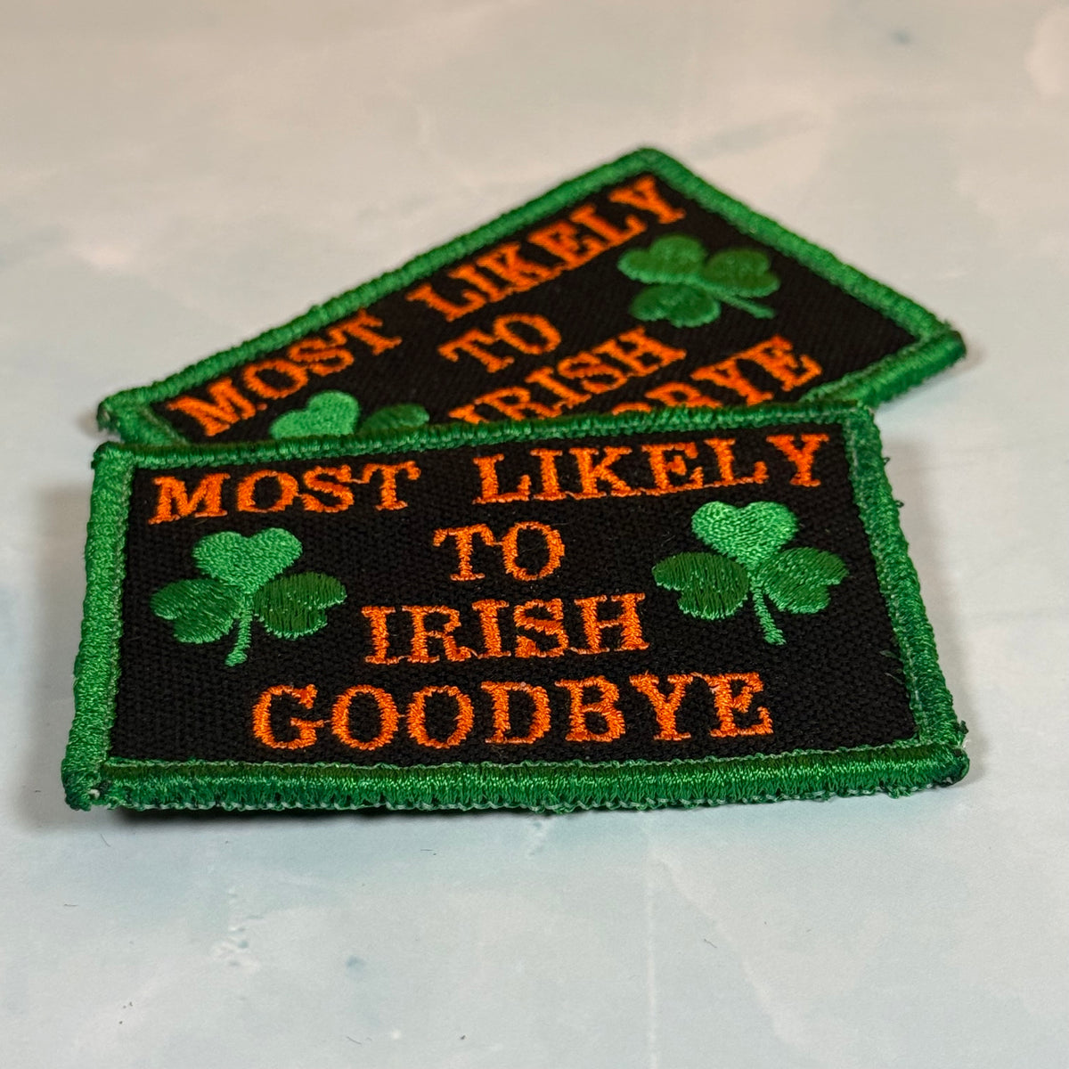 Two embroidered rectangular patches with green borders, green shamrocks, and orange text reading “MOST LIKELY TO IRISH GOODBYE” on a light gray background.