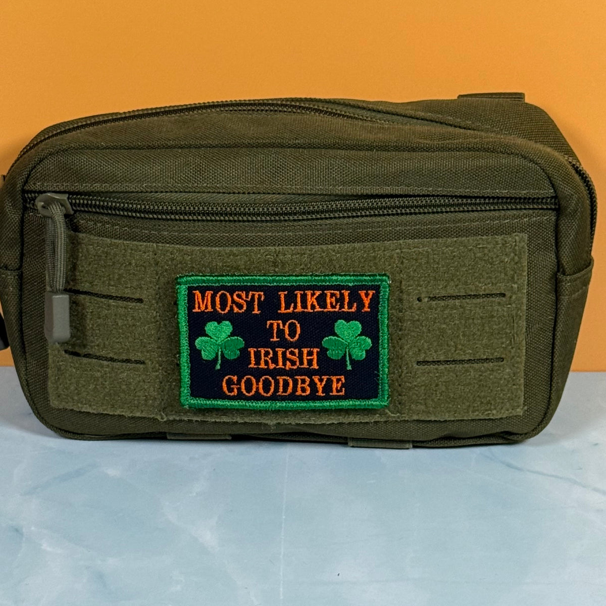 An olive green tactical pouch with a front zippered pocket and a rectangular embroidered patch that reads 'MOST LIKELY TO IRISH GOODBYE' in orange text with green shamrocks, set against an orange and light blue background.
