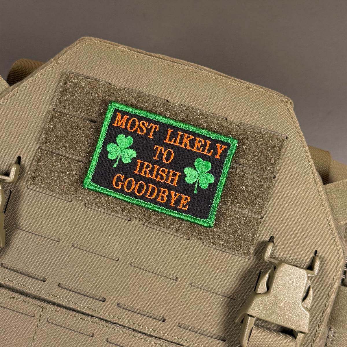 A rectangular embroidered patch with the text 'MOST LIKELY TO IRISH GOODBYE' in orange lettering on a black background, bordered in green and decorated with two green shamrocks, shown attached to a tan tactical vest.