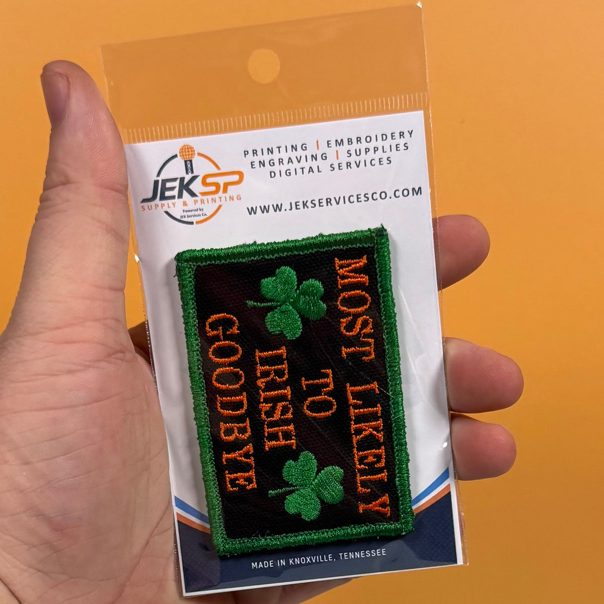 A hand holds a packaged embroidered patch against an orange background. The patch reads 'MOST LIKELY TO IREISH GOODBYE' in orange lettering with three green shamrocks on a black background, and it is mounted on a white card from Jeksp Service & Supply Co.