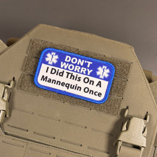 A rectangular embroidered patch with a blue border and white center reading “DON'T WORRY I Did This On A Mannequin Once,” attached to tan tactical gear with velcro and plastic buckles.