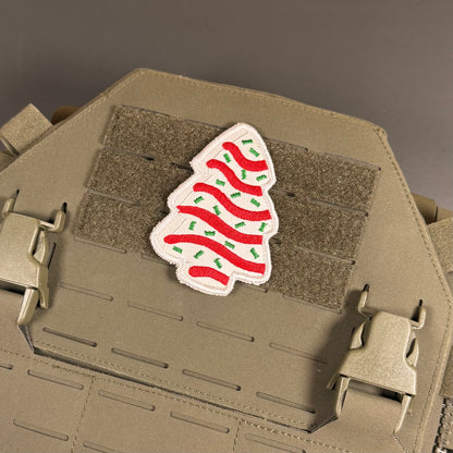 A Christmas tree-shaped embroidered patch with red stripes and green ornaments on a white background, attached to a tan tactical vest with MOLLE webbing and quick-release buckles.