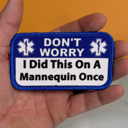I Did This On a Mannequin Once printed canvas morale patch held in a hand, featuring a blue border and a white center with the design letteringT WORRY I Did This On A Mannequin Once' and two white medical-style star symbols.