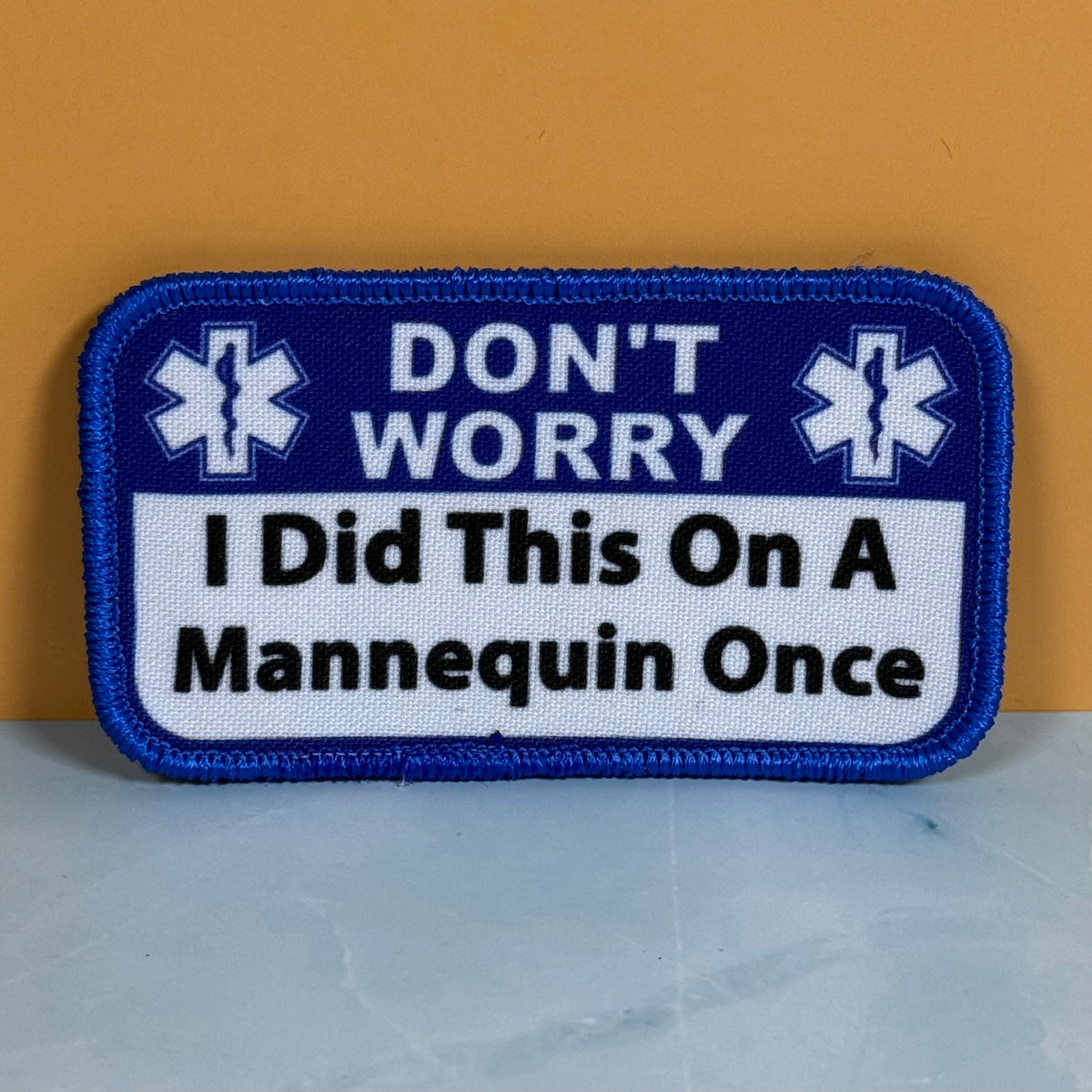 I Did This On a Mannequin Once printed canvas morale patch with rounded corners featuring the design letteringT WORRY I Did This On A Mannequin Once' in white and black on a blue and white background, placed against a mustard.