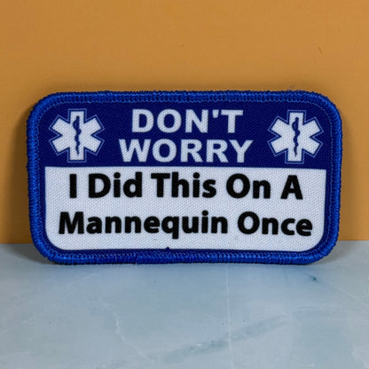 I Did This On a Mannequin Once printed canvas morale patch with rounded corners featuring the design letteringT WORRY I Did This On A Mannequin Once' in white and black on a blue and white background, placed against a mustard.