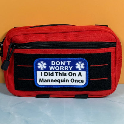I Did This On a Mannequin Once printed canvas morale patch, a red and black medical first aid pouch with a humorous patch that the design letteringT WORRY I Did This On A Mannequin Once' on a yellow and white background.
