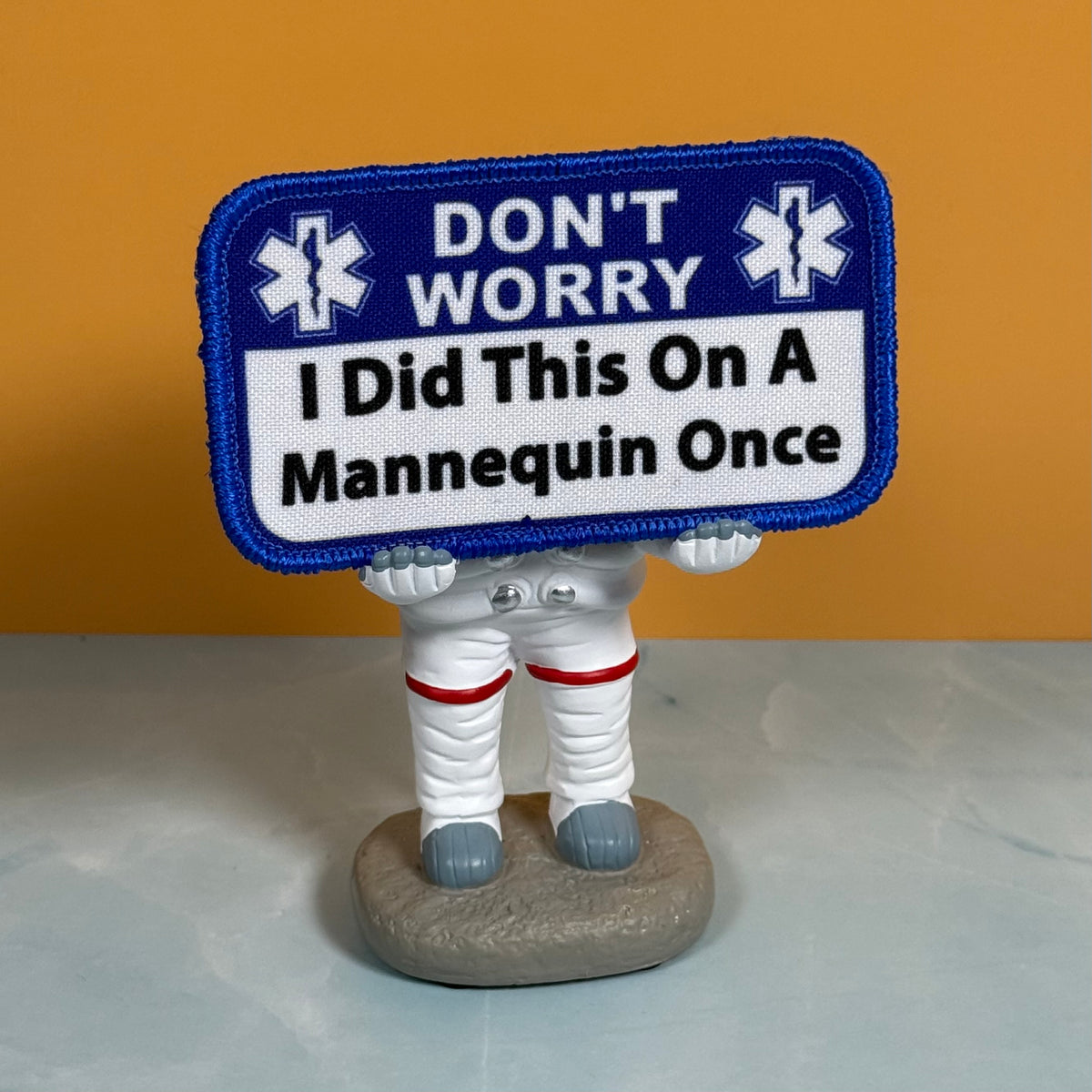 I Did This On a Mannequin Once printed canvas morale patch, a small astronaut figurine holding a blue and white printed patch that the design letteringT WORRY I Did This On A Mannequin Once,” set against an orange background.
