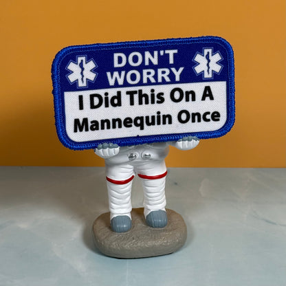 I Did This On a Mannequin Once printed canvas morale patch, a small astronaut figurine holding a blue and white printed patch that the design letteringT WORRY I Did This On A Mannequin Once,” set against an orange background.