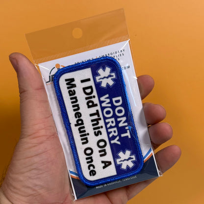 I Did This On a Mannequin Once printed canvas morale patch, a hand holds a packaged printed patch against a solid orange background. The patch is blue and white with the design lettering and 'DON'T WORRY' with medical-style.