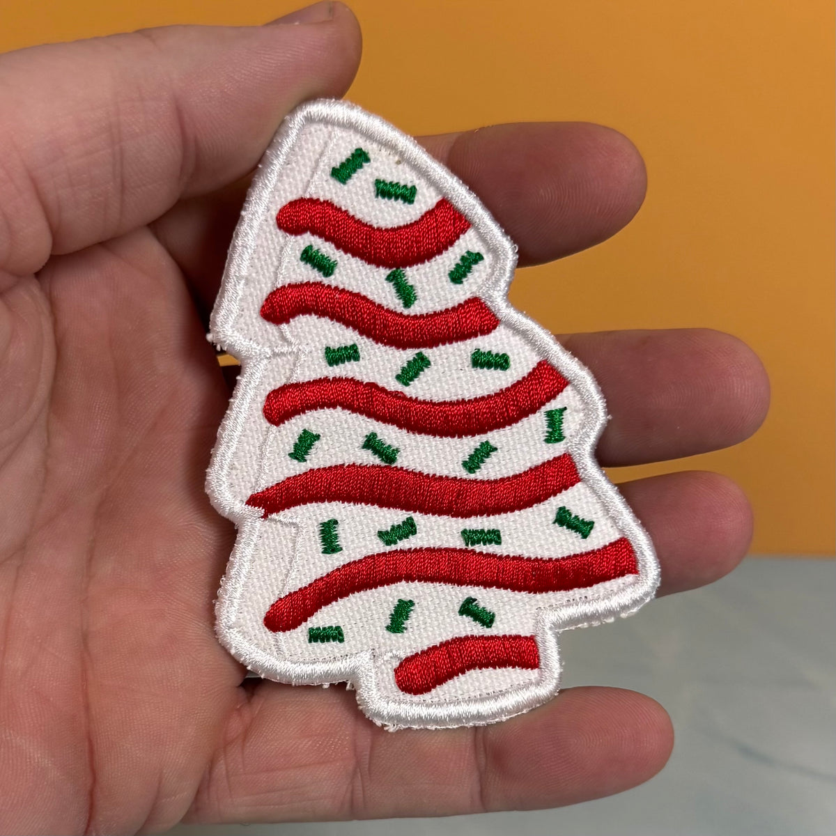An embroidered Christmas tree patch with red stripes and green accents, held in a person's hand against a solid orange background.