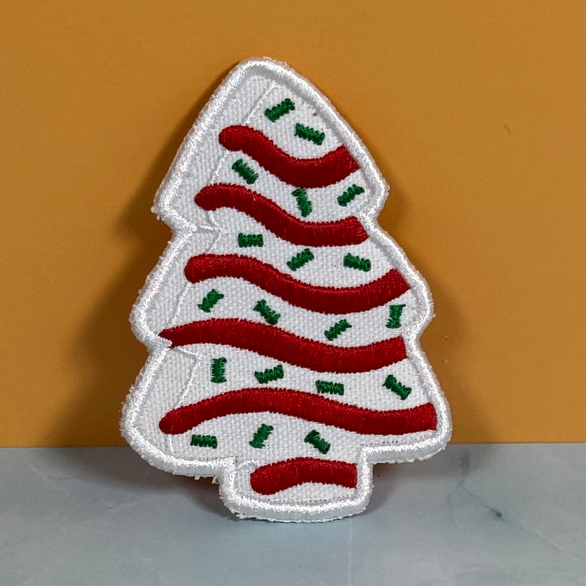 A Christmas tree-shaped embroidered patch with a white background, red wavy stripes, and small green accents, displayed against a mustard yellow background.