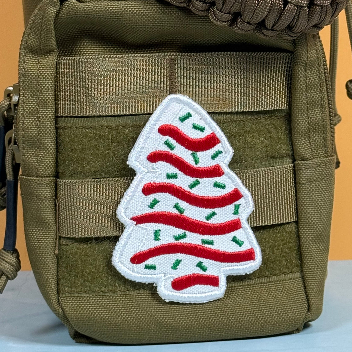 A white embroidered Christmas tree patch with red wavy stripes and green accents, attached to the side of an olive drab tactical pouch.
