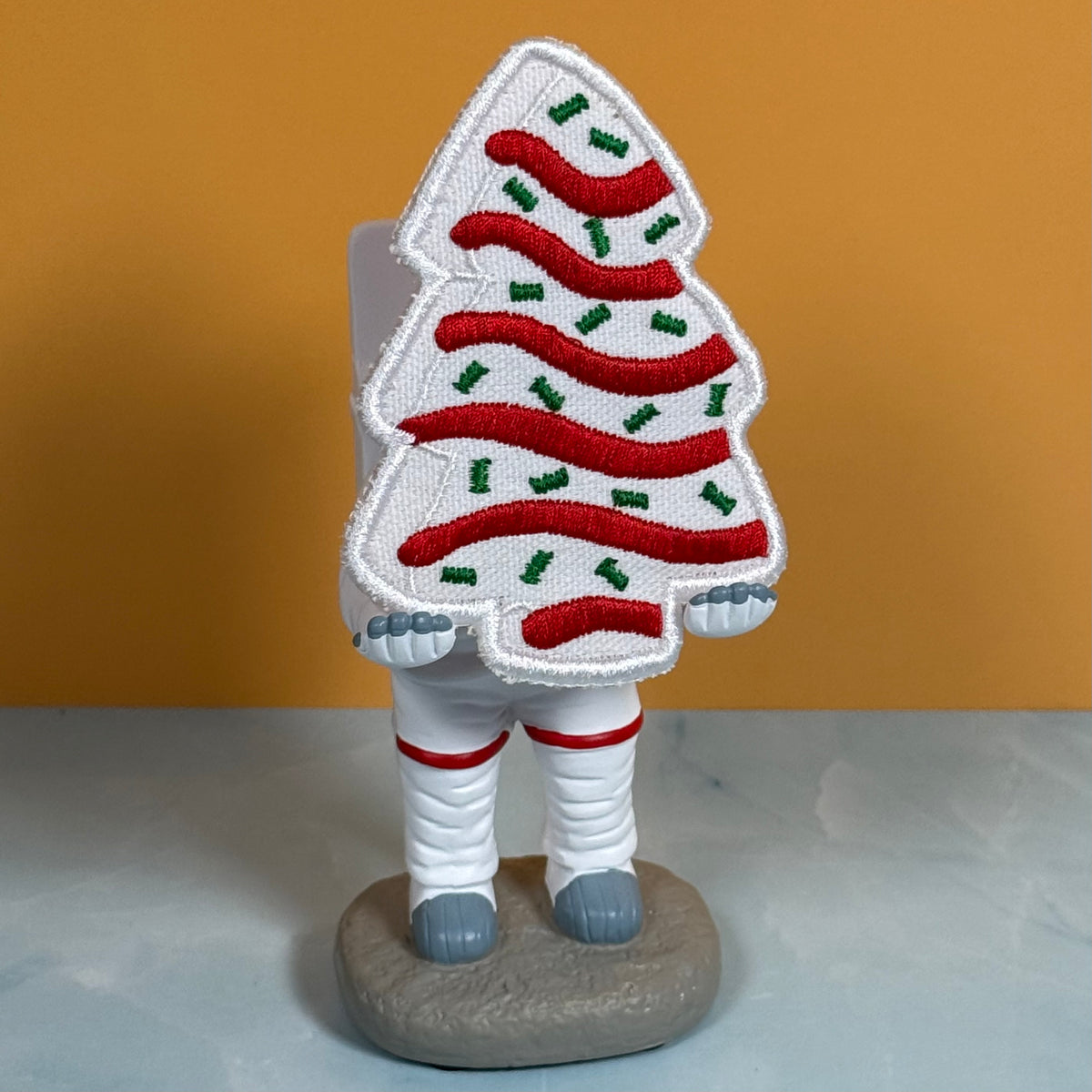 A decorative astronaut figurine with a Christmas tree-shaped helmet, featuring red and green embroidered stripes on a white background, standing on a gray base against a mustard yellow backdrop.