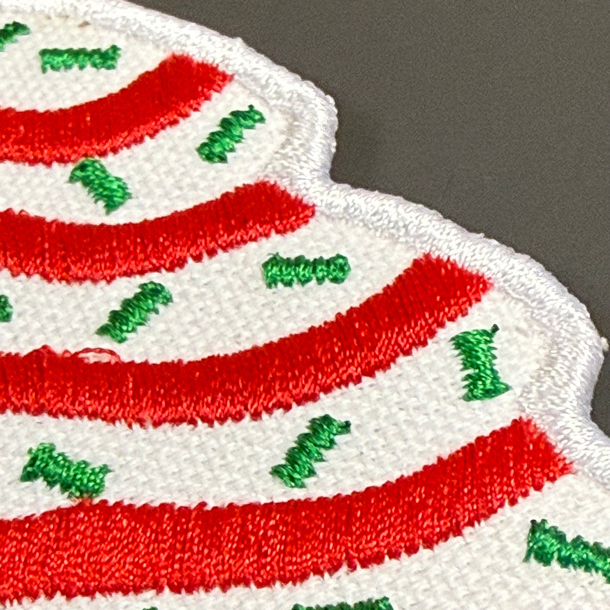 A close-up of a white embroidered patch with bold red curved stripes and scattered green embroidered accents, shown against a dark gray background.