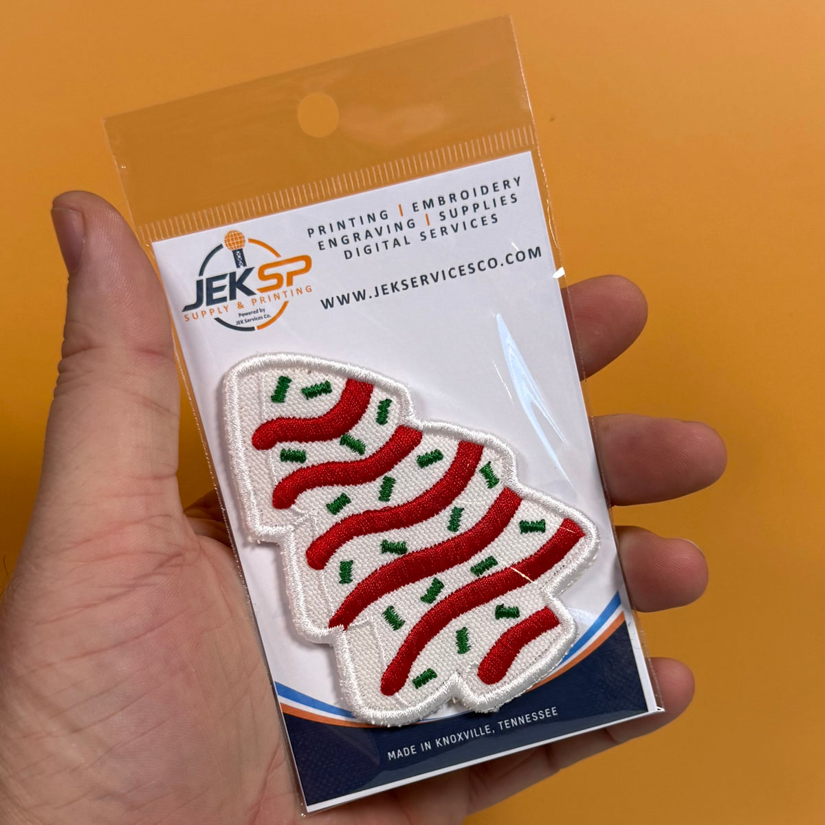 A hand holds a packaged embroidered patch shaped like a Christmas tree, featuring red stripes and green pine needles on a white background. The packaging shows the JEKSP branding against a solid orange background.