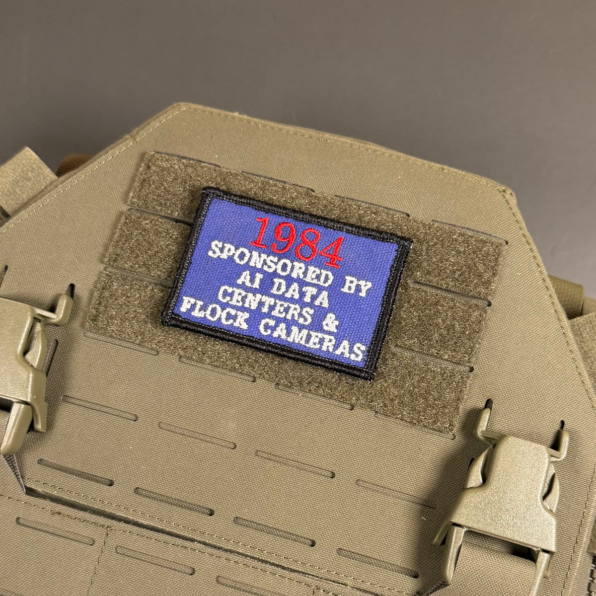 A close-up of a tactical plate carrier vest in coyote tan, featuring a rectangular blue patch on the front with red and white embroidered text reading '1984 SPONSORED BY AI DATA CENTERS & FLOCE CAMERAS'.
