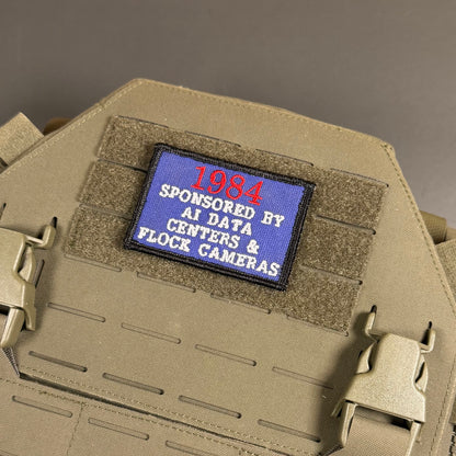 A close-up of a tactical plate carrier vest in coyote tan, featuring a rectangular blue patch on the front with red and white embroidered text reading '1984 SPONSORED BY AI DATA CENTERS & FLOCE CAMERAS'.