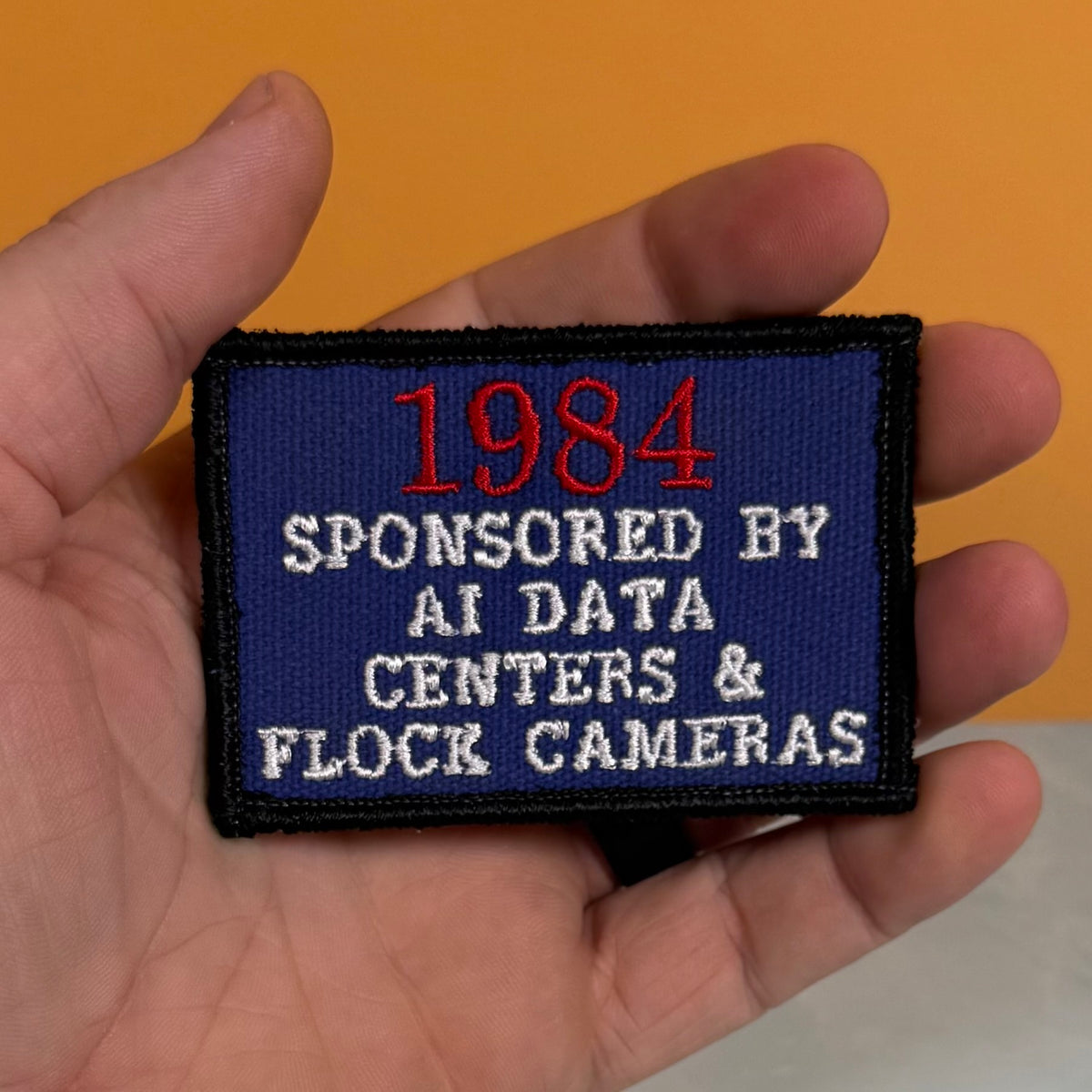 A hand holding a rectangular embroidered patch with a dark blue background, black border, red '1984' text, and white text reading 'SPONSORED BY AI DATA CENTERS & FLOCK CAMERAS' against a solid orange background.