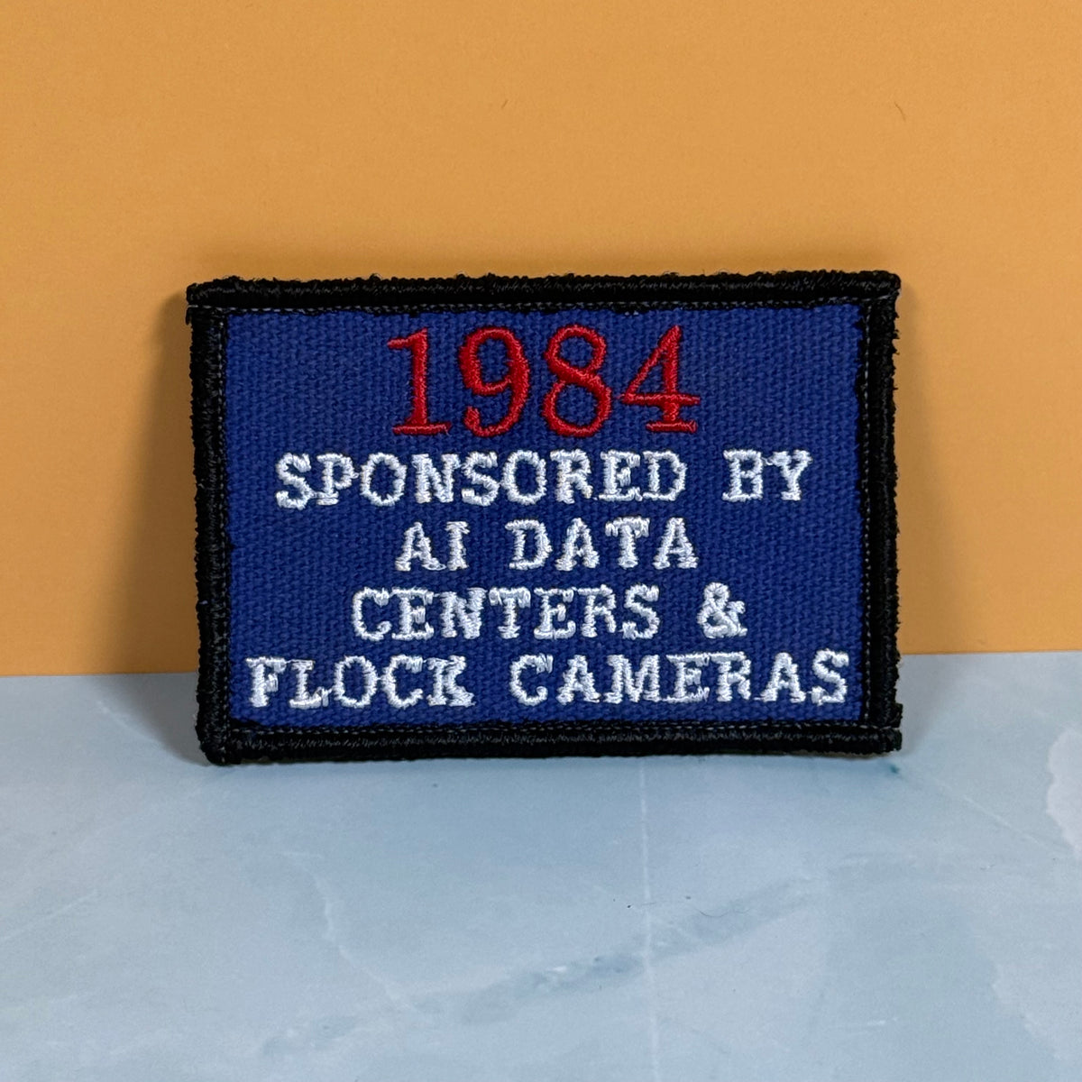 A rectangular embroidered patch with a blue field, black border, red '1984' text, and white text reading 'SPONSORED BY AI DATA CENTERS & FLOCK CAMERAS,' photographed against a two-tone orange and light gray background.