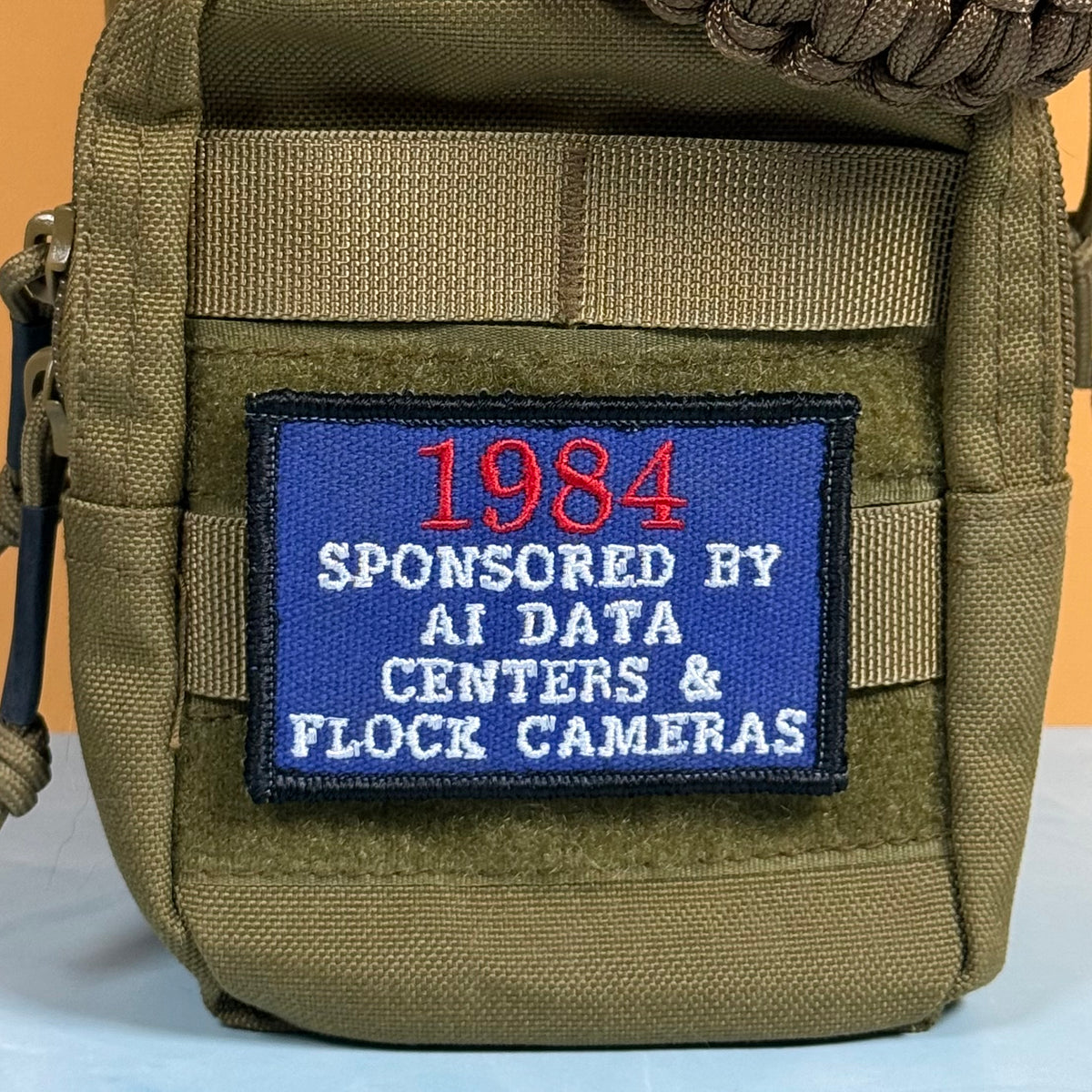 An olive green tactical pouch with a blue embroidered patch that reads '1984 SPONSORED BY AI DATA CENTERS & FLOCK CAMERAS' in red and white text.