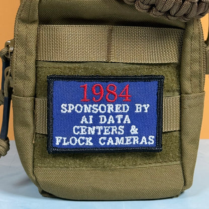 An olive green tactical pouch with a blue embroidered patch that reads '1984 SPONSORED BY AI DATA CENTERS & FLOCK CAMERAS' in red and white text.