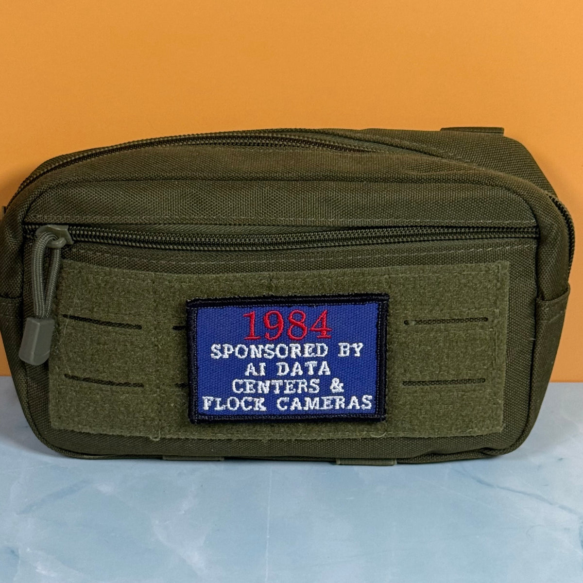 An olive green tactical pouch with a front zipper and a blue embroidered patch that reads '1984 SPONSORED BY AI DATA CENTERS & FLOCK CAMERAS', set against an orange background and light blue surface.