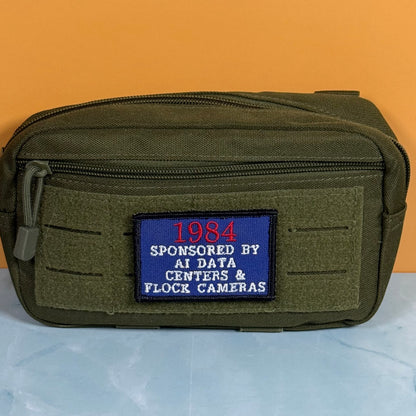 An olive green tactical pouch with a front zipper and a blue embroidered patch that reads '1984 SPONSORED BY AI DATA CENTERS & FLOCK CAMERAS', set against an orange background and light blue surface.