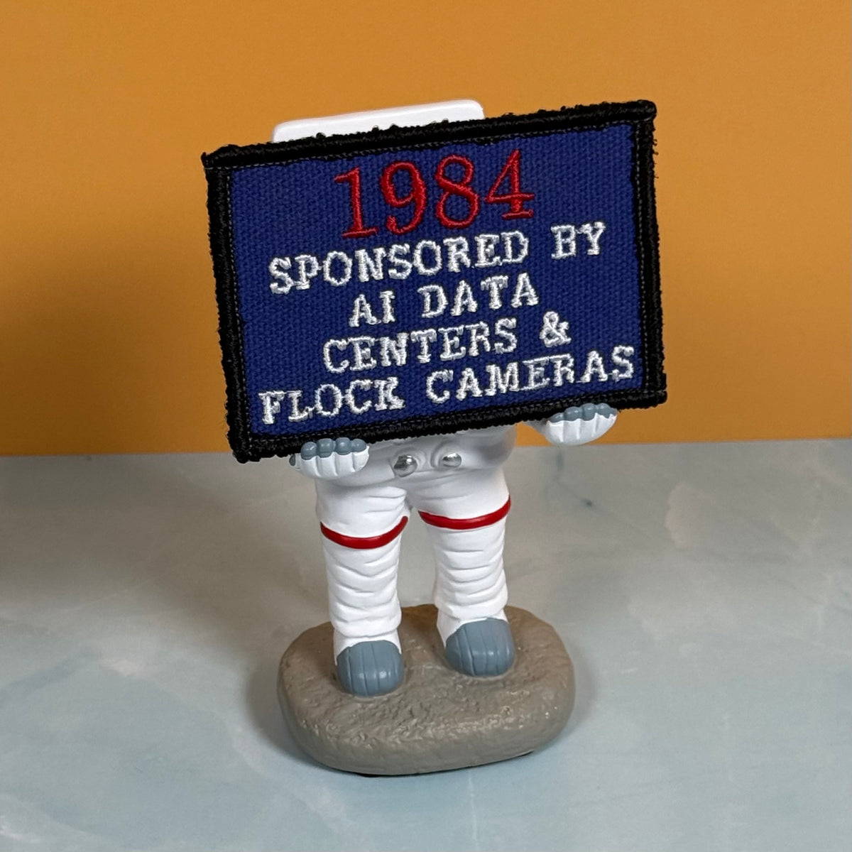 A small astronaut figurine holding a rectangular embroidered sign that reads “1984 SPONSORED BY AI DATA CENTERS & FLOCK CAMERAS.” The astronaut is white with red stripes and gray boots and base, photographed against a warm orange wall and pale gray surface.