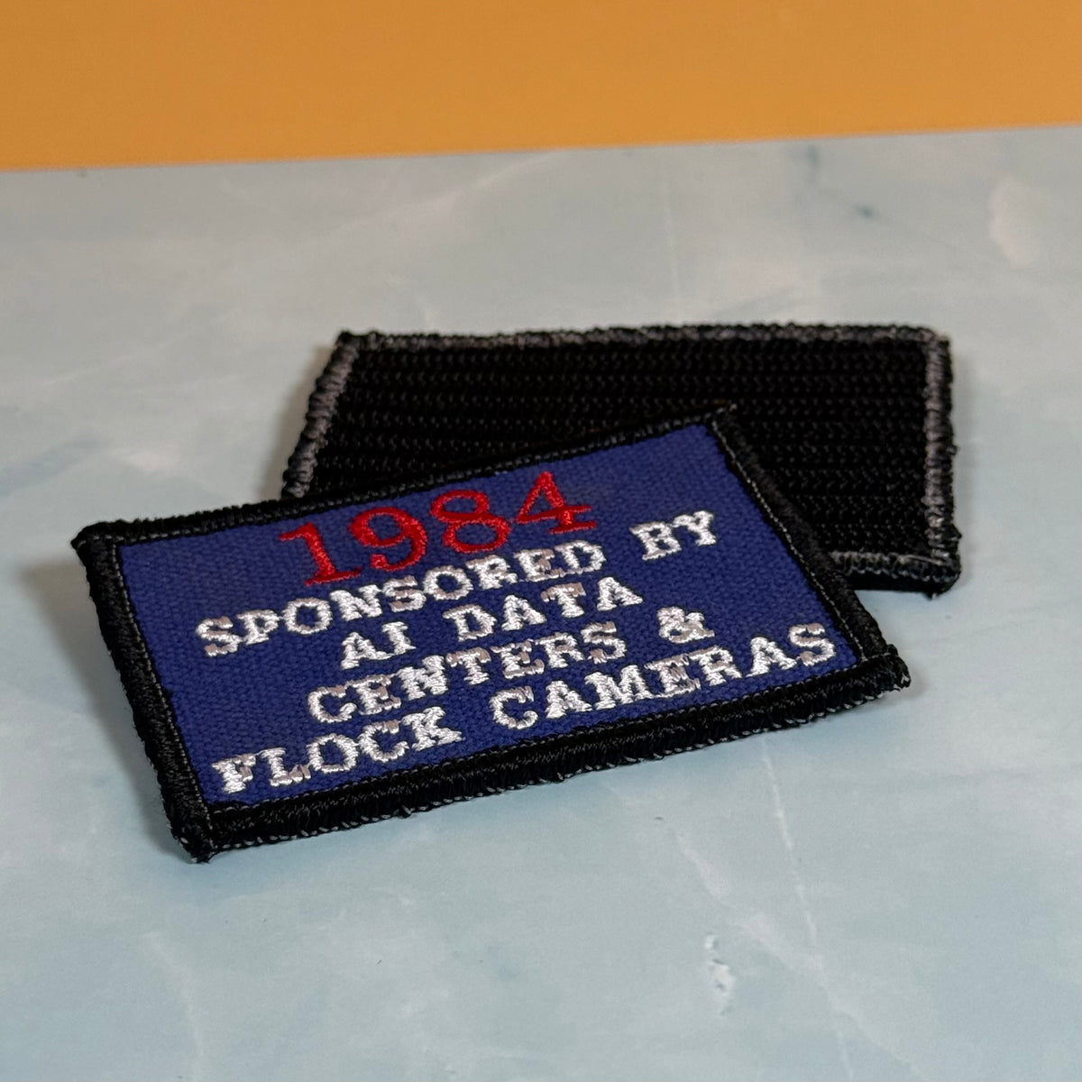 Two embroidered patches on a light marble-like surface with an orange background. The front patch is blue with red and white text reading '1984 SPONSORED BY AI DATA CENTERS & FLOCK CAMERAS' and has a black border.