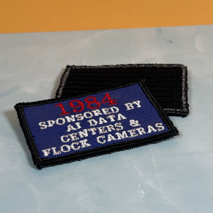 Two embroidered patches on a light marble-like surface with an orange background. The front patch is blue with red and white text reading '1984 SPONSORED BY AI DATA CENTERS & FLOCK CAMERAS' and has a black border.