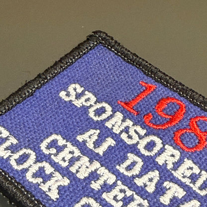 Close-up of a rectangular embroidered patch with a blue field, black stitched border, white text reading 'SPONSORED BY AI DATA', and red '198' text, shown against a neutral taupe background.