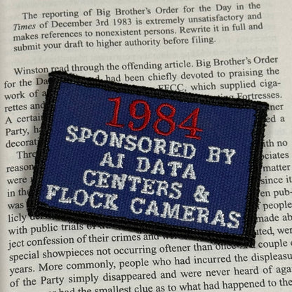 A rectangular embroidered patch with a dark blue background and black stitched border, featuring the text '1984 SPONSORED BY AI DATA CENTERS & FLOCC CAMERAS' in red and white lettering, placed on top of printed text.