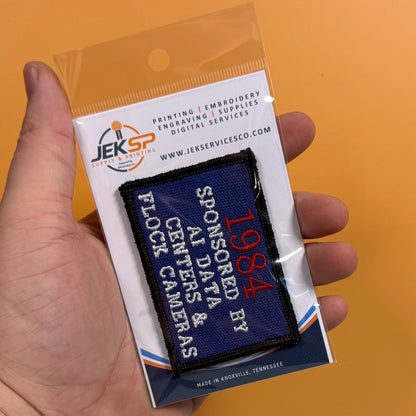 A hand holds a packaged embroidered patch against a solid orange background. The patch is dark blue with red and white text reading '1984 SPONSORED BY AI DATA & BLOCK CAMERAS'.