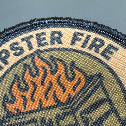 Close-up of a woven embroidered patch with the words "DESPERATE FIRE" in dark blue lettering on a beige background, above an orange and olive green flame graphic.