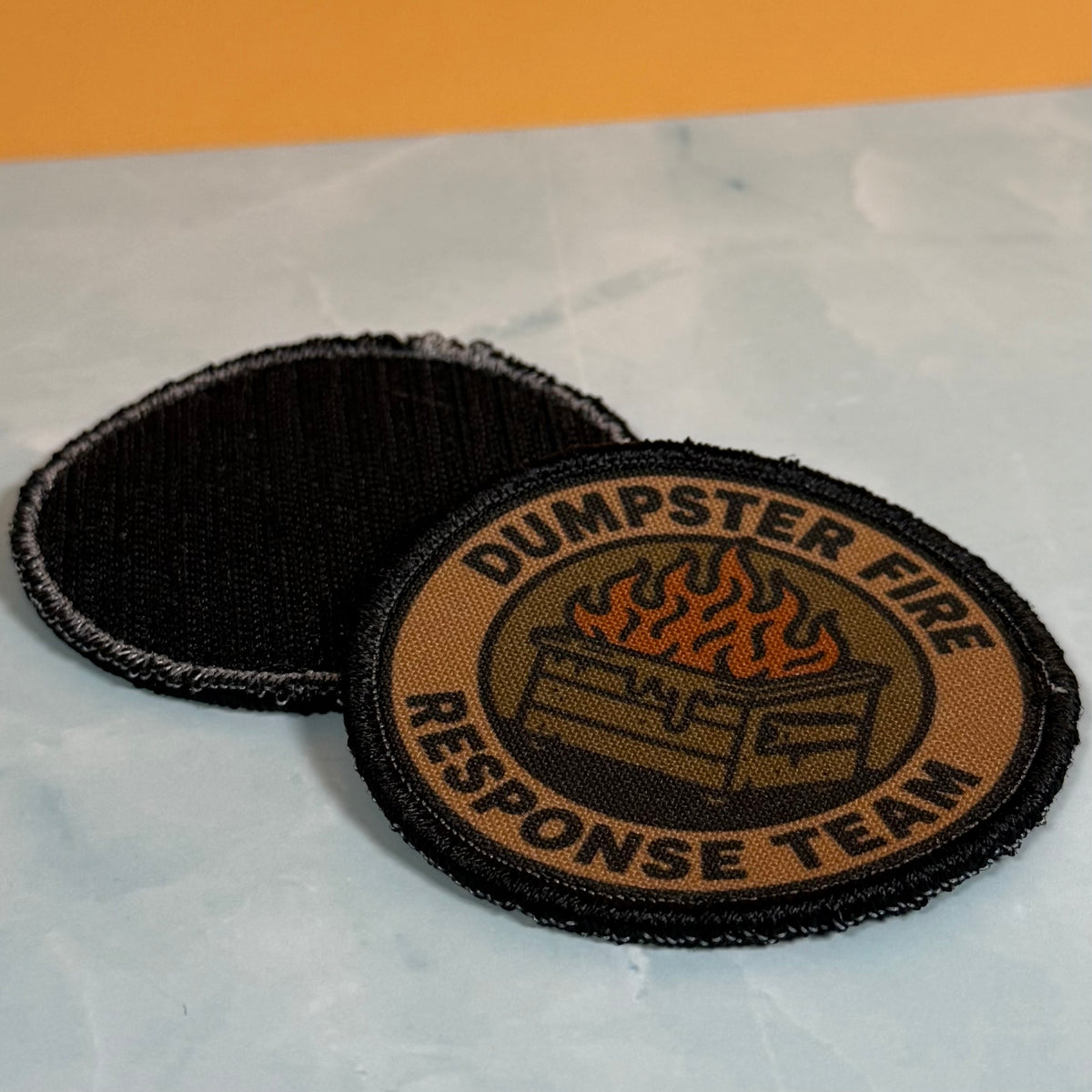 Two circular embroidered patches for the Dumpster Fire Response Team, one showing the front design with a burning dumpster and flames, and the other showing the black reverse side, placed on a light marble surface with an orange background.